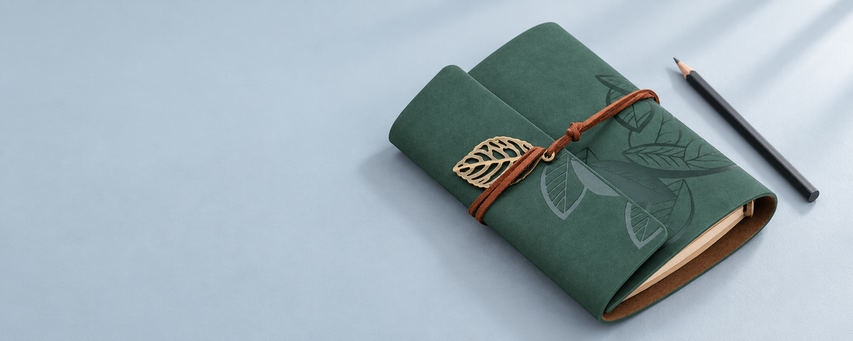 Product styling illustration of the green leaf-cover journal on a light blue-gray desk